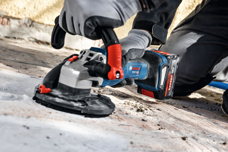 Eine Person mit Sicherheitsausrüstung verwendet einen Akku-Betonschleifer auf einer rauen Oberfläche. Anwendungsbild