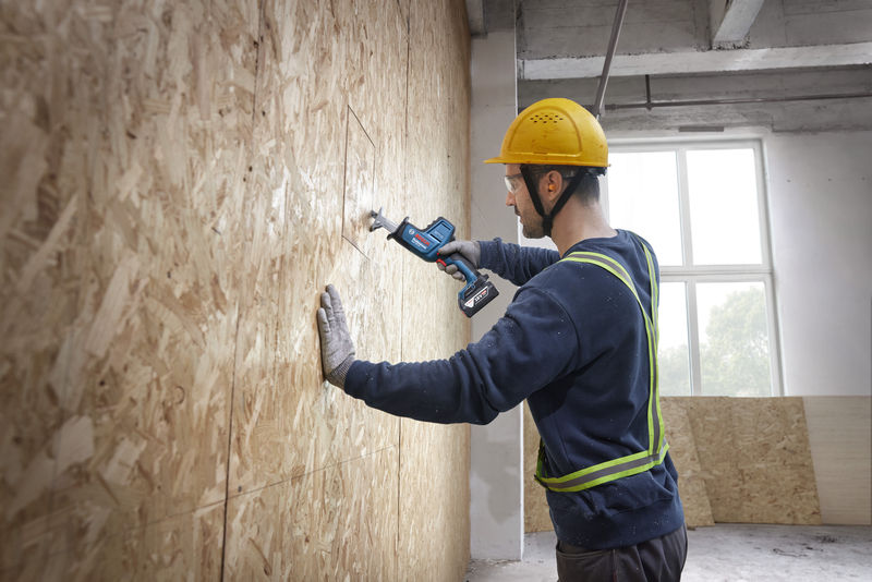 Eine Person mit Sicherheitsausrüstung schneidet mit einer Säbelsäge in eine Holzwand. Anwendungsbild