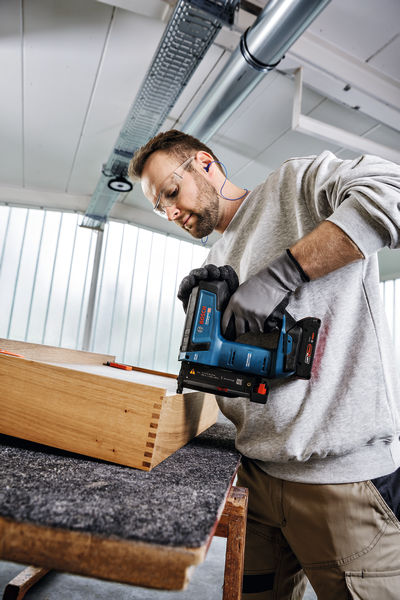 Eine Person mit Sicherheitsausrüstung befestigt Holzverbindungen mit einem Akku-Nagler. Anwendungsbild