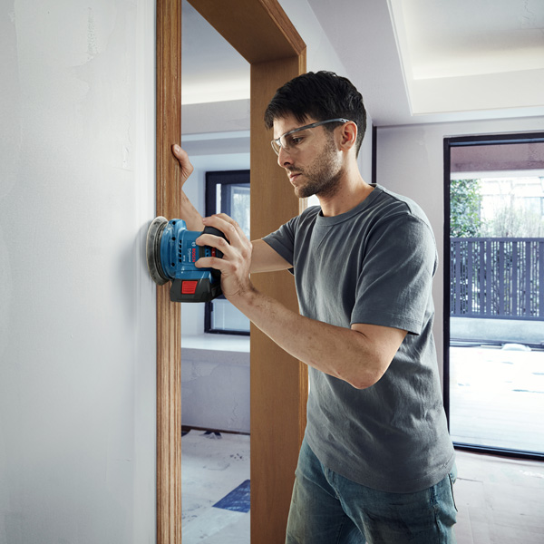 Eine Person mit Schutzausrüstung schleift eine Wand mit einem Akku-Schleifgerät. Anwendungsbild