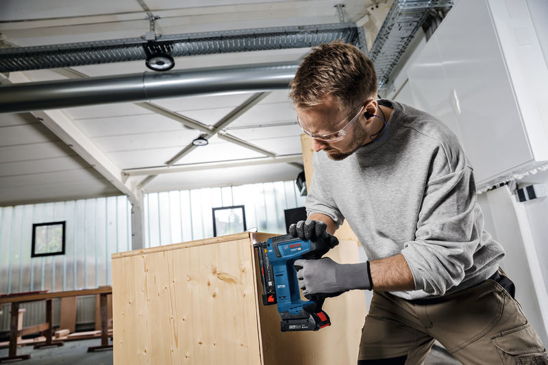 Eine Person mit Sicherheitsausrüstung heftet in einer Werkstatt Holzplatten zusammen. Anwendungsbild