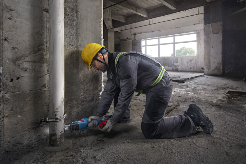 Eine Person mit Sicherheitsausrüstung schneidet mit einer Säbelsäge ein Rohr in der Nähe einer Betonwand durch. Anwendungsbild