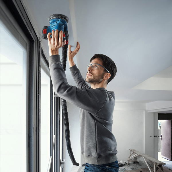 Eine Person mit Schutzausrüstung schleift mit einem elektrischen Schleifgerät eine Decke in der Nähe eines Fensters. Anwendungsbild
