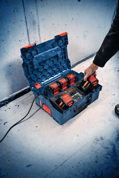 Eine Person legt eine Batterie in ein blaues Mehrfach-Ladegehäuse auf einem Betonboden. Anwendungsbild