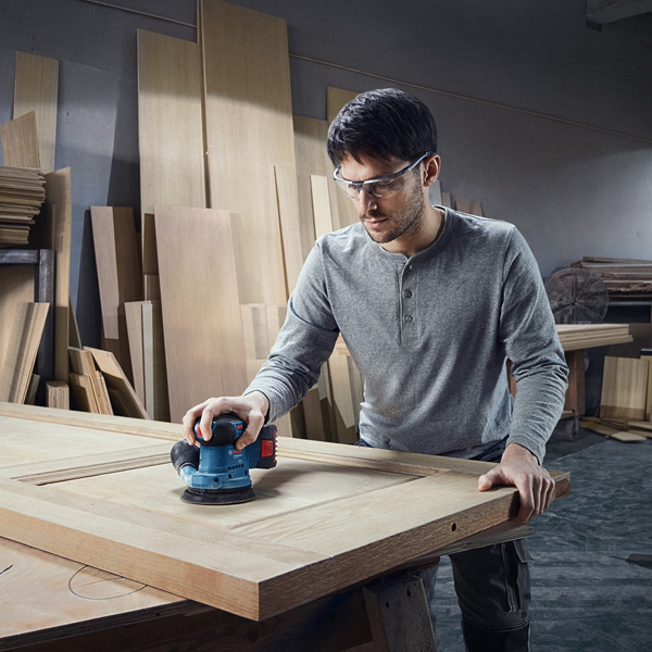 Eine Person mit Schutzausrüstung schleift eine Holztür mit einem elektrischen Schleifgerät. Anwendungsbild