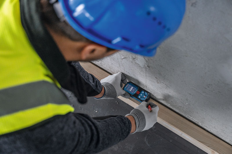 Eine Person mit Sicherheitsausrüstung verwendet ein Lasermessgerät an einer Wand. Anwendungsbild
