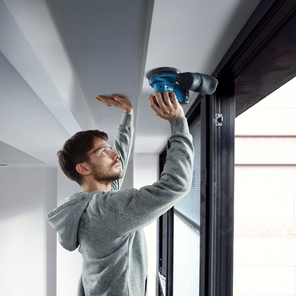Eine Person mit Schutzausrüstung schleift mit einem elektrischen Schleifgerät eine Deckenkante. Anwendungsbild