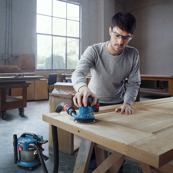 Eine Person mit Schutzausrüstung schleift eine Holztür mit einem angeschlossenen Exzenterschleifer. Anwendungsbild