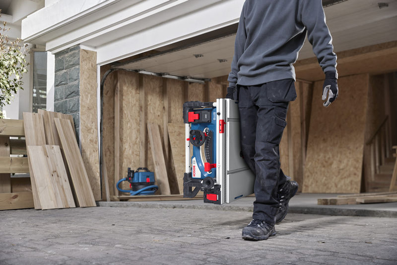 Eine Person mit Sicherheitsausrüstung trägt auf einer Baustelle eine tragbare Tischkreissäge. Anwendungsbild Nahaufnahme